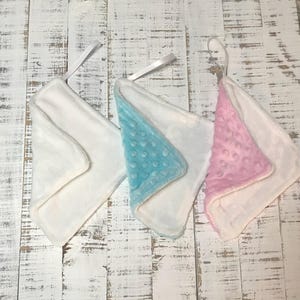 May include: Three white baby burp cloths with satin ribbon loops. One cloth has a light blue minky dot corner, and another has a pink minky dot corner. The cloths are folded into triangles.