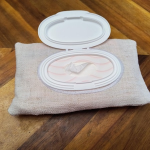 Puede incluir: Un recipiente de tela beige para toallitas con una tapa de pl&aacute;stico blanco. La tapa est&aacute; abierta, revelando una pila de toallitas. El recipiente es rectangular con bordes redondeados. El fondo es una superficie de madera.