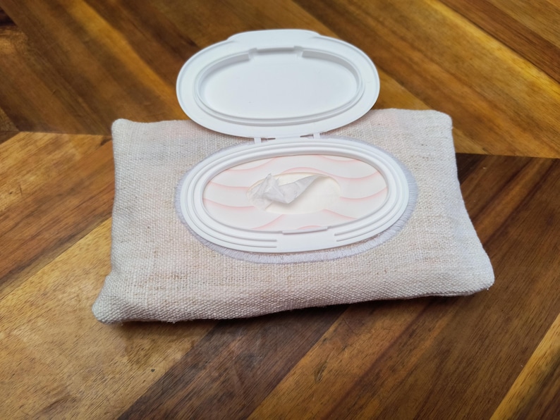 Puede incluir: Un recipiente de tela beige para toallitas con una tapa de pl&aacute;stico blanco. La tapa est&aacute; abierta, revelando una pila de toallitas. El recipiente es rectangular con bordes redondeados. El fondo es una superficie de madera.