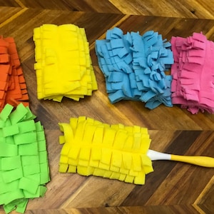 May include: A selection of colourful cleaning pads in orange, yellow, blue, pink, and green. A yellow duster with a white handle and yellow grip is also shown. The items are arranged on a wooden surface.