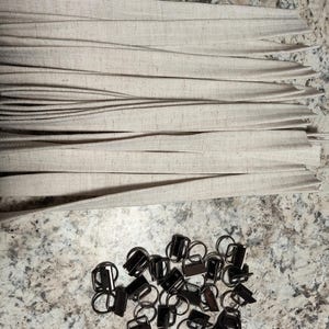 May include: A pile of black metal slider buckles with D-rings. The buckles are arranged in a random pattern on a light gray surface. There is a stack of beige fabric strips in the background.