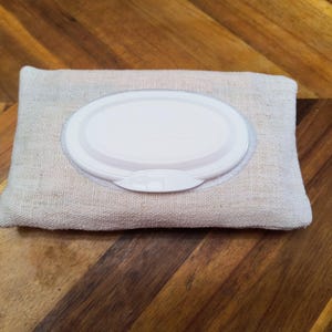 Puede incluir: Un dispensador de toallitas de tela beige rectangular con una tapa blanca de forma ovalada. La tapa tiene una peque&ntilde;a abertura curva para dispensar las toallitas. El dispensador est&aacute; sobre un fondo de madera.