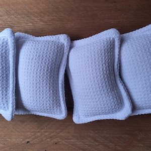 Puede incluir: Cuatro almohadas rectangulares blancas con una superficie texturizada y punteada y un borde liso y redondeado. Las almohadas están dispuestas sobre una superficie de madera, mostrando su diseño limpio y sencillo. Las almohadas son de un blanco claro y brillante.