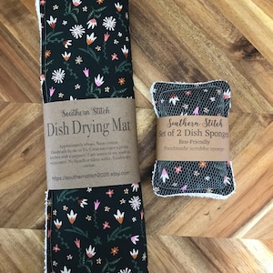 May include: A dark green dish drying mat and two dish sponges, both with a floral design. The mat features white, pink, and orange flowers. The sponges are eco-friendly and handmade. The text on the mat reads "Southern Stitch Dish Drying Mat".