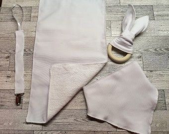 Puede incluir: Un conjunto de tres artículos para bebés blancos: un babero, un babero y un mordedor con un anillo de madera. El babero y el babero están hechos de un tejido suave y absorbente. El mordedor tiene forma de conejo y tiene una superficie suave y texturizada.