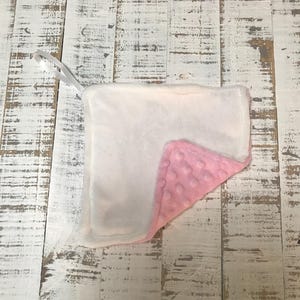 以下が含まれることがあります： 白とピンクのベビーブランケット。吊り下げ用のループが付いています。ブランケットは柔らかく、豪華な素材で作られています。ピンクの面は、テクスチャード加工されたドット柄です。