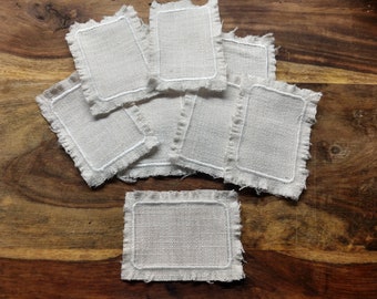 Puede incluir: Un juego de ocho posavasos de lino blanco con borde deshilachado y borde blanco cosido. Los posavasos están apilados sobre una superficie de madera.