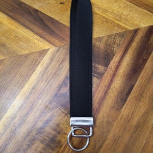 Könnte beinhalten: Ein schwarzes Handgelenkband mit einem silberfarbenen Metallverschluss und einem Spaltring. Das Band besteht aus einem glatten, einfarbigen schwarzen Material. Der Hintergrund ist eine Holzoberfläche mit einem braunen und beigen Muster.