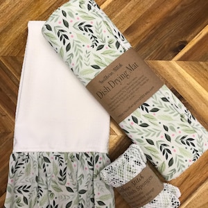 May include: A set of three kitchen items in a leafy green and pink pattern. The set includes a dish drying mat, a dish towel with a ruffled edge, and two dish sponges. The dish drying mat and dish towel are made of fabric, and the dish sponges are made of a mesh material. The packaging for the items is brown paper with white text that reads "Southern Stitch Dish Drying Mat" and "Southern Stitch 2 Dish Sponges".
