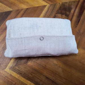 Puede incluir: Una bolsa rectangular de tela blanquecina con una solapa asegurada por un broche plateado. La bolsa est&aacute; hecha de un material texturizado y natural y descansa sobre una superficie de madera con un patr&oacute;n de espiga.