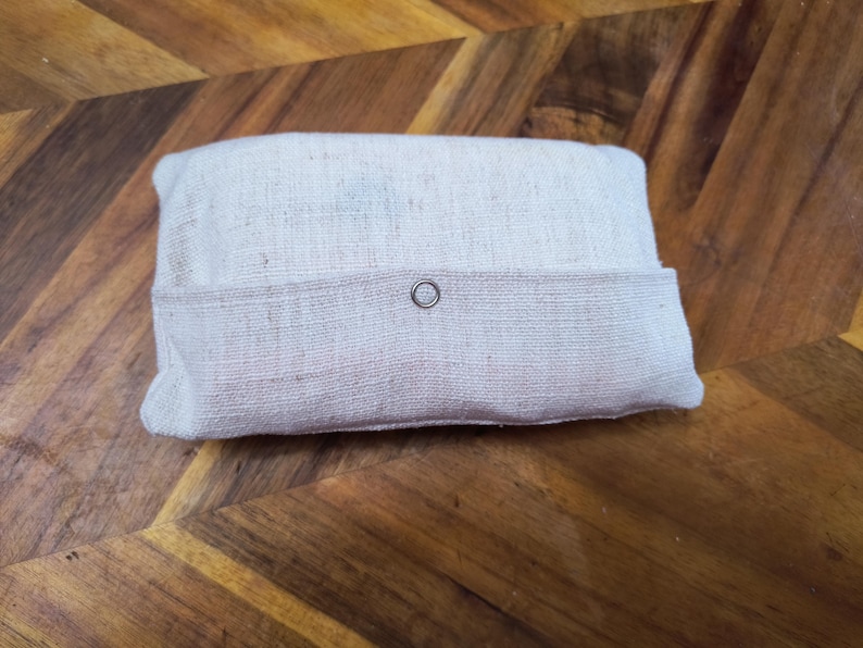 Puede incluir: Una bolsa rectangular de tela blanquecina con una solapa asegurada por un broche plateado. La bolsa est&aacute; hecha de un material texturizado y natural y descansa sobre una superficie de madera con un patr&oacute;n de espiga.