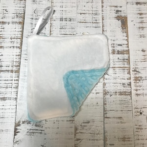 Puede incluir: Una manta para bebé blanca y azul claro con un lazo de cinta de raso. La manta tiene un centro blanco suave y afelpado y un borde azul claro con lunares. La manta es rectangular y parece estar hecha de un material suave.