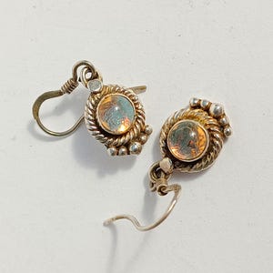 May include: A pair of silver earrings with iridescent, round, amber-colored stones. The stones are set in a twisted rope design, surrounded by small silver beads. The earrings have a hook closure.