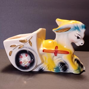 May include: A ceramic donkey-shaped planter pulling a cart. The donkey is white with yellow and blue accents and black painted eyes. The cart is white with a black wheel featuring red spokes and gold leaf branch detailing on the side.