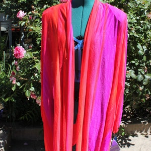 May include: A vibrant, flowing, open-front garment in shades of red and fuchsia, displayed on a dress form. The fabric appears lightweight and sheer. The background features a lush green bush with pink roses.