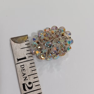 May include: A cluster of iridescent, clear crystal beads forms a decorative brooch. The brooch is oval-shaped and features a variety of crystal sizes. A measuring tape is next to the brooch. The brooch is approximately 1 inch in diameter.