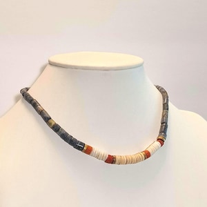 May include: A beaded necklace featuring a sequence of dark grey cylindrical beads, followed by segments of small, flat, cream-colored disc beads interspersed with thin, vibrant orange and metallic gold-toned spacer beads along the center.
