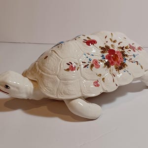 May include: A ceramic turtle figurine with a white body and a shell decorated with red, pink, and blue floral patterns. The turtle has a detailed head and legs, and the shell has a textured appearance.