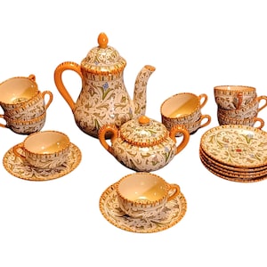 May include: A ceramic tea set featuring a teapot, a lidded sugar bowl, and multiple cups and saucers. Each piece is decorated with an intricate green and brown foliate pattern on a cream background, accented with solid orange trim and handles.