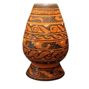 May include: A ceramic vase with a tapered neck and a wide, rounded body resting on a flared pedestal base. The surface features alternating bands of dark brown and terracotta orange, decorated with intricate geometric patterns and wave motifs.