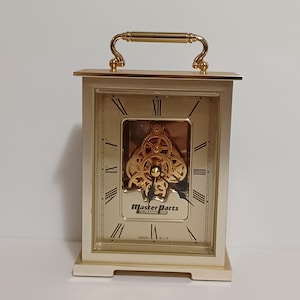 May include: A rectangular, off-white clock with a gold-colored handle and Roman numerals. The clock face has a gold-colored background with visible gears and the text "MASTER PARTS" and "HOWARD MILLER".