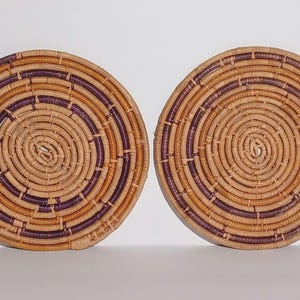 May include: Two circular woven wall baskets featuring a coiled design. The baskets are constructed from natural tan fibers interspersed with dark purple dyed coils, creating a repeating concentric pattern across the flat, round surfaces.