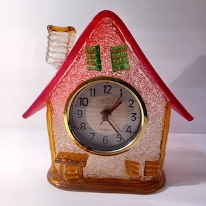 May include: A red and orange glass house shaped clock with a white face and black hands. The clock has a gold trim and says "Quartz" on the face.