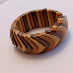 May include: A wooden bracelet with a geometric design. The bracelet features a series of rectangular and triangular wooden pieces in shades of brown, tan, and beige. The bracelet is flexible and appears to be made of multiple connected segments.