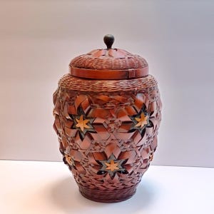 May include: A handcrafted, brown woven basket with a lid and a dark wooden knob. The basket features a repeating star pattern with accents of yellow and green. The basket is a decorative storage container.