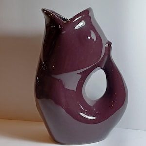 May include: A glossy, plum-colored ceramic pitcher with a unique, organic shape. The pitcher features a large, circular opening on the side and a flared top. The design is modern and sculptural, suitable for decorative use.