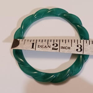 May include: A circular green bangle bracelet with a twisted, rope-like texture. A white measuring tape is positioned across the center of the bracelet, showing an inner diameter of approximately 2.5 inches for this solid, smooth-surfaced accessory.
