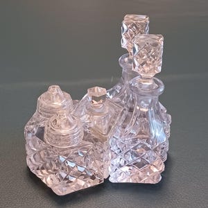 May include: A clear, cut-glass vanity set with multiple bottles and jars. The set includes two perfume bottles with square stoppers, two small jars with domed lids, and a small, square bottle. The glass has a diamond pattern.
