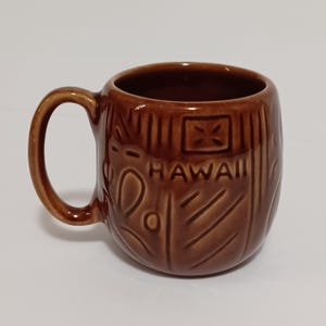 May include: Brown ceramic mug with a handle, featuring embossed tribal designs and the word "HAWAII". The mug has a glossy finish and a cylindrical shape, perfect for coffee or tea.