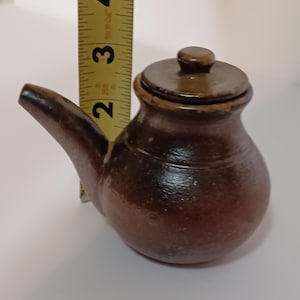 May include: A small, brown ceramic teapot with a lid and a spout. The teapot is approximately 2 inches tall. The teapot has a rounded body and a dark brown color. The lid is a lighter brown color.