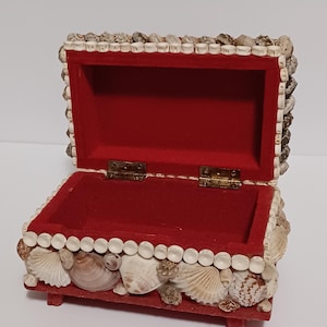 May include: Open red velvet-lined jewelry box decorated with seashells. The box is rectangular with a hinged lid and small wooden feet. The exterior is adorned with various sizes and shapes of seashells.