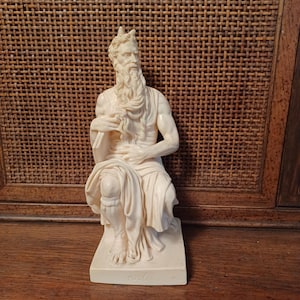 May include: A cream-colored figurine of a seated, bearded figure with small horns on the head, wearing draped robes. The figure holds a tablet in one hand and rests the other on the lap. The base of the statue features the embossed text MOSÈ.