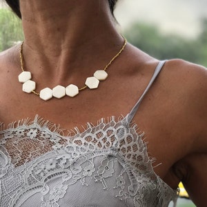 May include: A light gray camisole with lace detailing and spaghetti straps. A gold chain necklace with white hexagonal pendants is worn. The image is taken outdoors with a blurred green background.