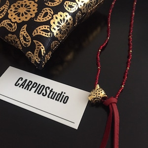 May include: A red beaded necklace with a gold-colored leopard head pendant and red leather tassels. A black and gold patterned pillow is in the background. The business card reads CARPIOstudio.