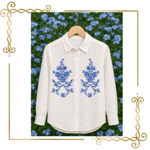 Könnte beinhalten: Ein langärmeliges weißes Hemd mit Knopfleiste und zwei symmetrischen blauen Blumenstickereien auf den Vorderteilen. Das Hemd hat einen klassischen Kragen und wird auf einem Holzkleiderbügel präsentiert, mit detaillierten blauen Mustern.