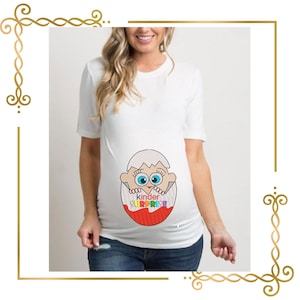 Peut inclure: Un t-shirt blanc à manches courtes arborant un motif de personnage de dessin animé sortant d'une coquille d'œuf fissurée. Le motif inclut le texte "kinder SURPRISE" en lettres colorées, avec la moitié inférieure de l'œuf en rouge vif.