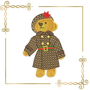 May include: An embroidered golden-yellow teddy bear wearing a brown and tan patterned trench coat with a double-breasted front, a green and red striped belt with a gold buckle, and a matching patterned beret topped with a small red leaf accent.