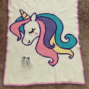 May include: A white crocheted baby blanket featuring a colorful unicorn design. The unicorn has a yellow horn, pink and purple mane, and teal accents. A pair of white baby shoes rests on the blanket.
