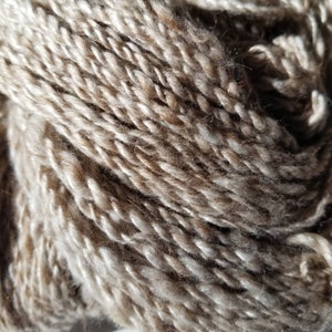 May include: A close-up of a skein of yarn in a light brown and beige color. The yarn is made of a blend of natural fibers, giving it a soft and textured look.