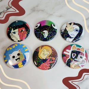 May include: Six round holographic buttons with different colourful designs. The buttons feature various characters and designs, including a cat, a person with a crown, and a person with a mask.