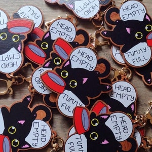 Puede incluir: Un conjunto de llaveros de madera con gatos negros que muestran diferentes lemas. Los llaveros tienen un diseño rojo y blanco con el texto "Cabeza: Vacía" y "Barriga: Llena".