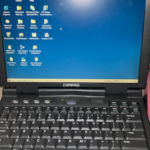 May include: A black Compaq laptop featuring a QWERTY keyboard and a screen displaying the Microsoft Windows 98 operating system desktop. The screen shows various software icons including Internet Explorer, Microsoft Outlook, and Recycle Bin.