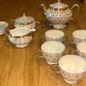 May include: A vintage tea set with a floral pattern in white, gold, and iridescent colors. The set includes a teapot, sugar bowl, creamer, six teacups, and a stack of saucers. The rims and handles are gold.