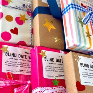 May include: A collection of rectangular craft kits wrapped in various papers including white with pink dots and smiley faces, brown kraft paper, and multicolored stripes. Each package features a white label reading BLIND DATE WITH YOUR NEXT FAVORITE HOBBY.