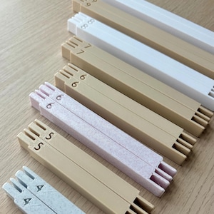 May include: An assortment of rectangular, interlocking wooden pieces in shades of white, beige, and pink. Each piece is marked with a number from 4 to 10 and features slots. The pieces are arranged on a light brown surface. Measurements are in cm.
