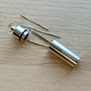 May include: A cylindrical silver metal needle case with a screw-top lid and a gold-colored keyring. The word Needles is engraved on the side of the case. Several thin, metallic sewing needles are positioned inside and outside of the open container.